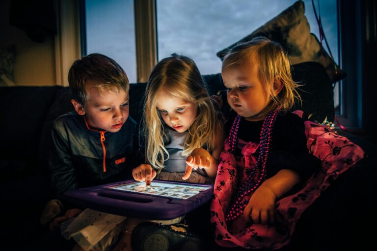 Three young children engrossed in a tablet while sitting indoors, illuminated by the screen.