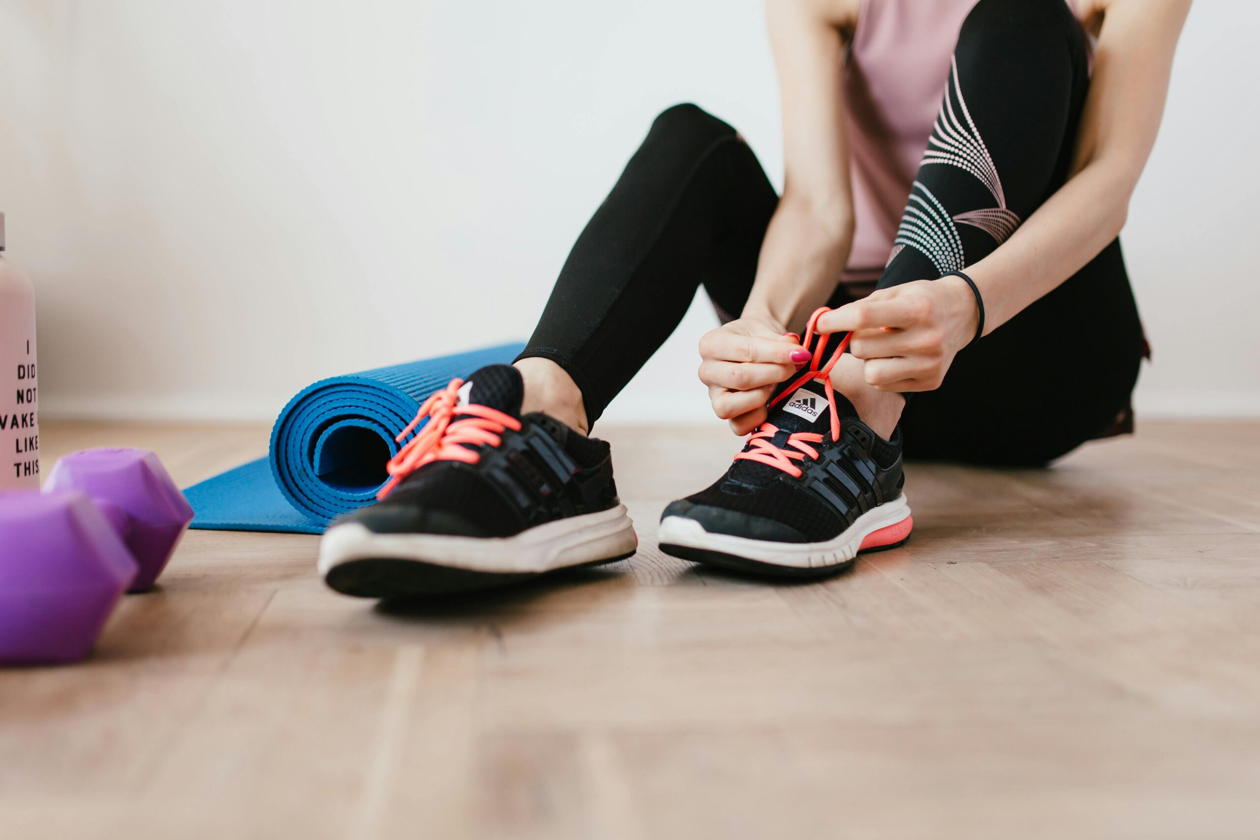 Fit woman tying shoelaces, preparing for indoor workout with yoga mat and dumbbells.