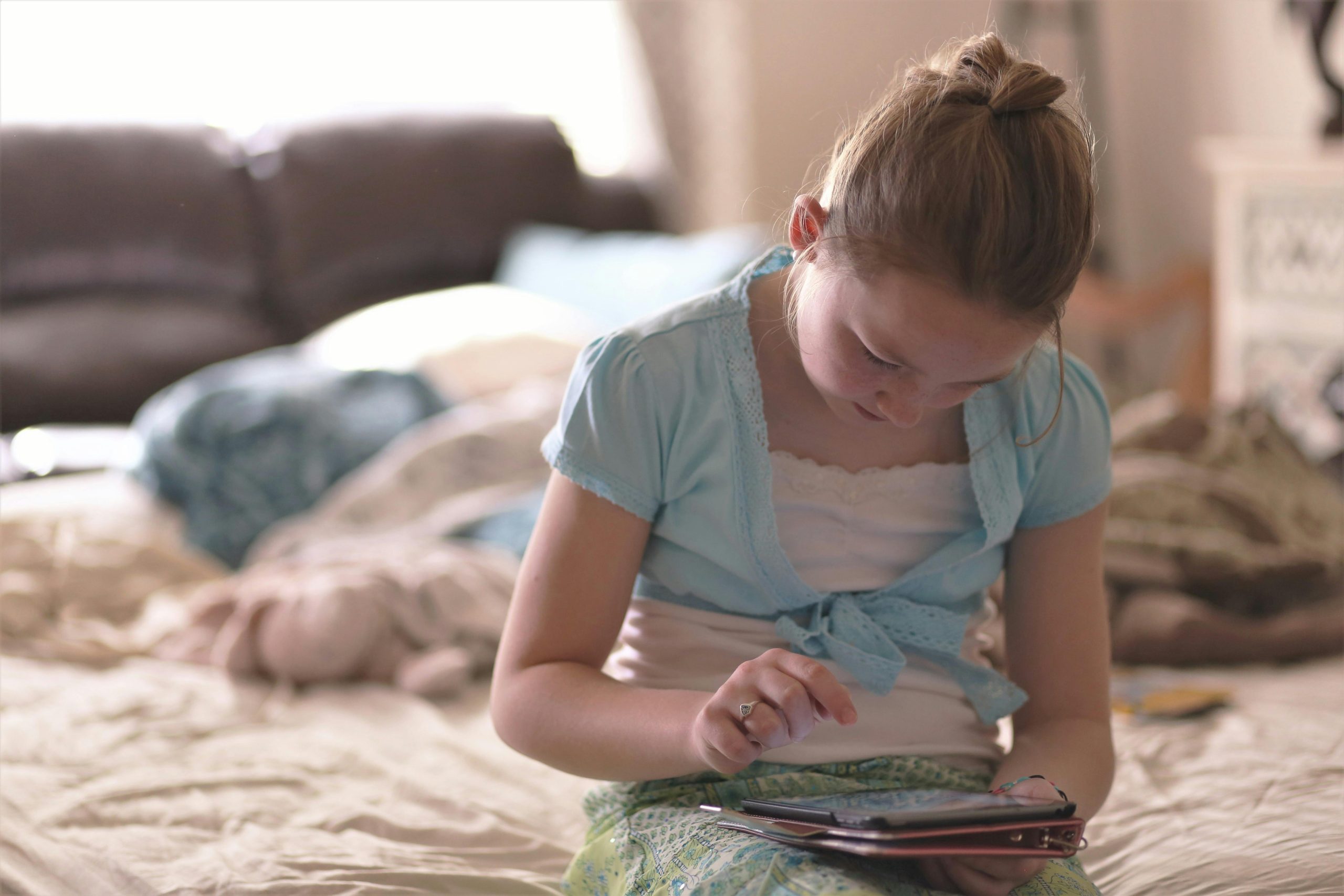 A young girl sitting on a bed, focused on using a tablet device in a cozy indoor setting.