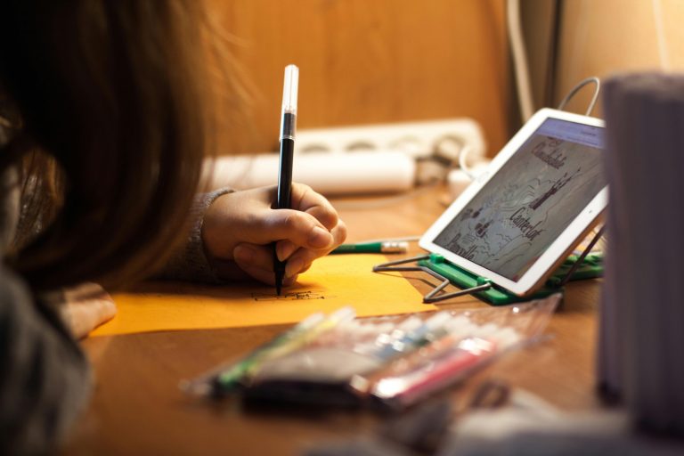 A young girl drawing at a desk with a tablet for reference, highlighting creative use of technology.